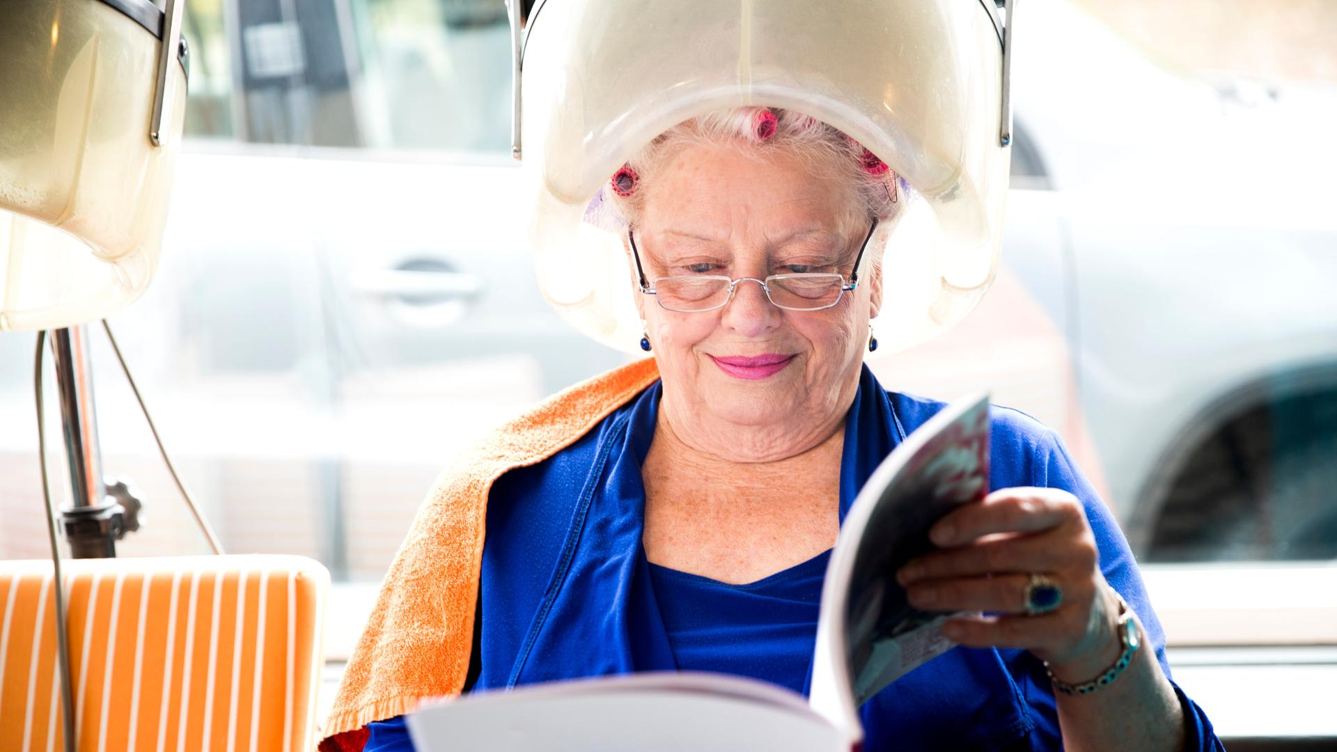 Eine ältere Frau sitzt lächelt unter einer Trockenhaube und liest dabei eine Zeitschrift.