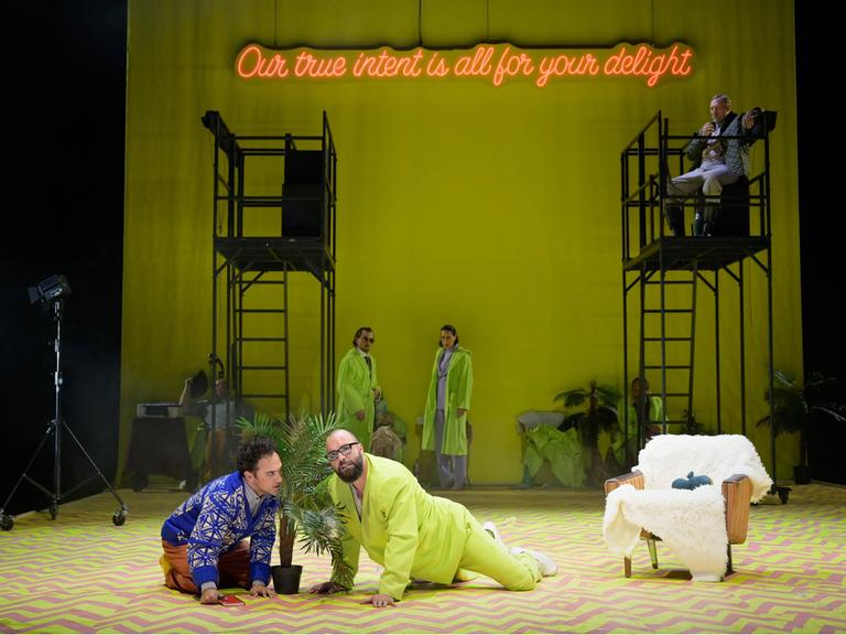 Auf einer Theaterbühne sind zwei Männer zu sehen. Sie befinden sich auf allen Vieren; zwischen ihnen steht eine Topfpflanze. 