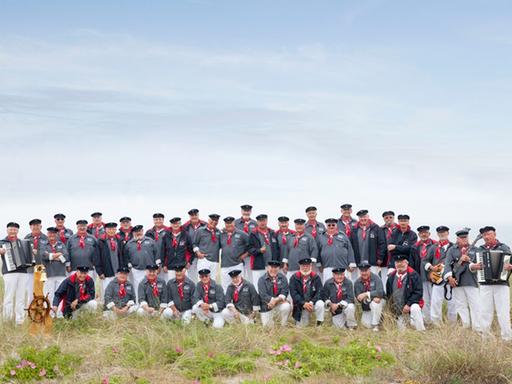 Grupenbild des Chores in den Dünen. Alle Männer tragen ein gestreiftes blau-weißes Hemd, einen Hut, ein rotes Halstuch und weiße Hosen.