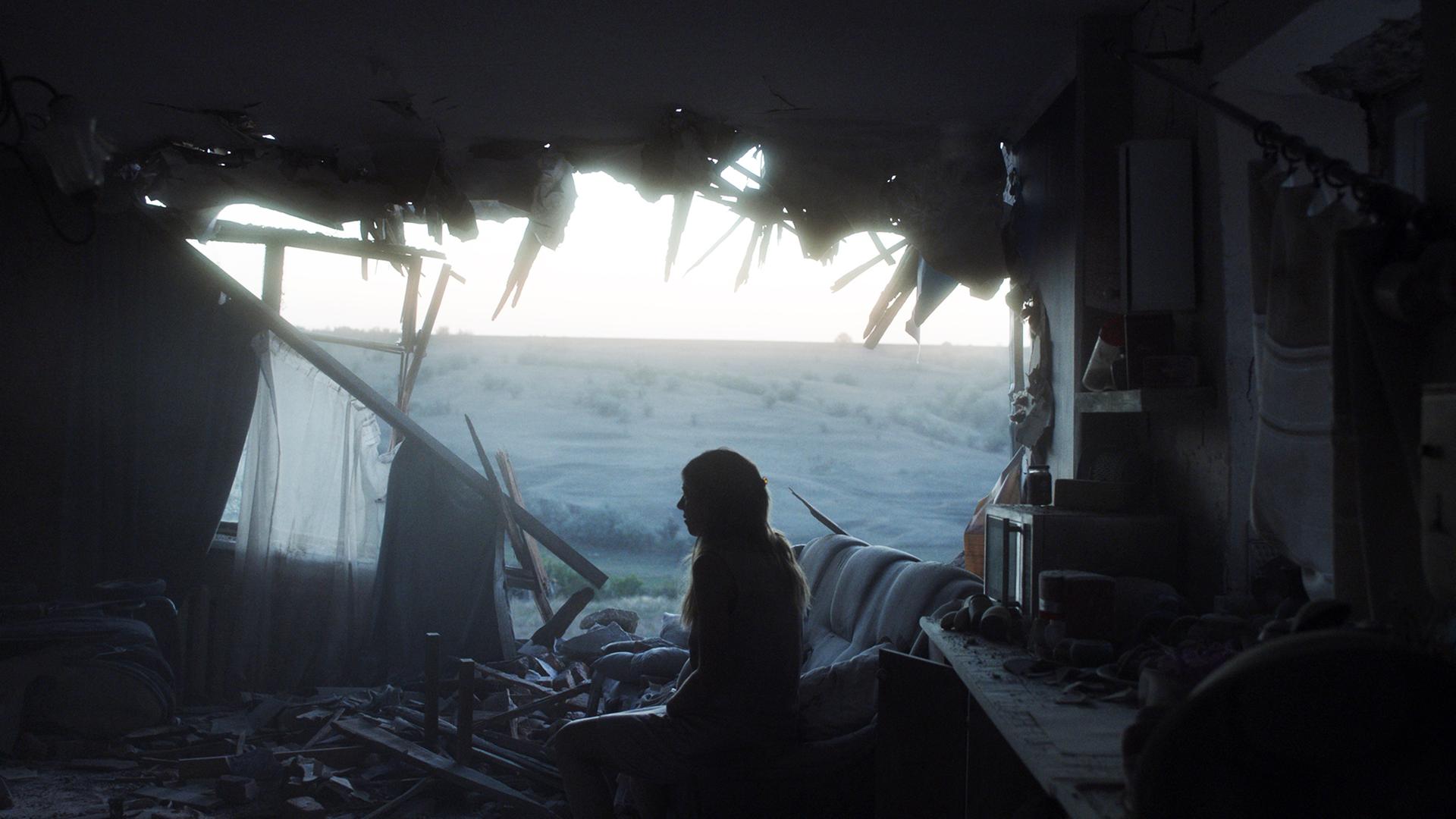 Eine junge Frau sitzt in einem zerstörtem Wohnzimmer und schaut durch eine zerstörte Hauswand nach draußen.