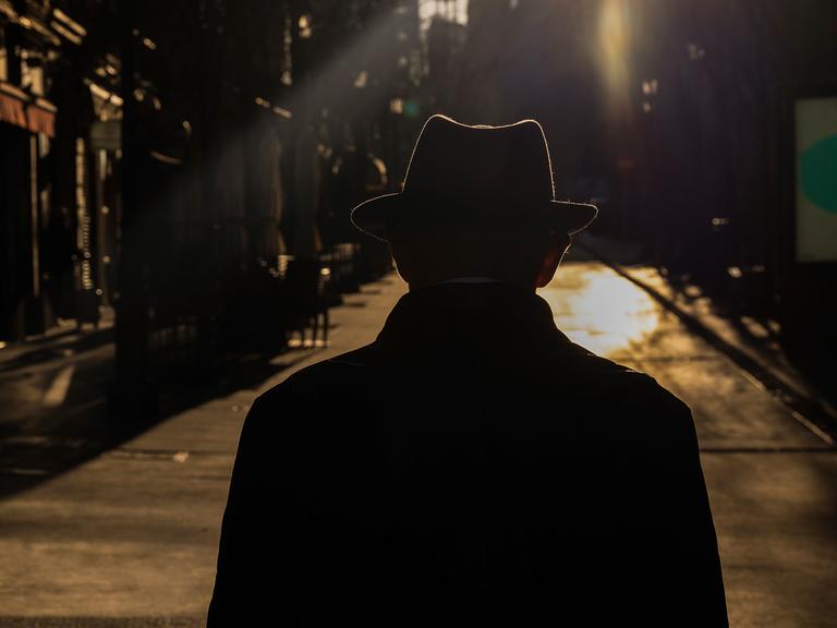Ein Mann mit Hut geht mit dem Rücken zur Kamera eine Straße in der Dunkelheit entlang. 