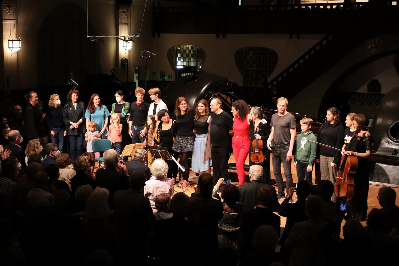 Musikerinnen und Musiker sowie einige Kinder stehen in legerer Kleidung aufgereiht auf einer Bühne. Umrahmt werden sie von Maschinen im Jugendstil-Kraftwerk Heimbach (Eifel) sowie Zuhörerinnen und Zuhörern, die ihnen Applaus spenden. Musikerinnen und Musiker sowie einige Kinder stehen in legerer Kleidung aufgereiht auf einer Bühne. Umrahmt werden sie von Maschinen im Jugendstil-Kraftwerk Heimbach (Eifel) sowie Zuhörerinnen und Zuhörern, die ihnen Applaus spenden.