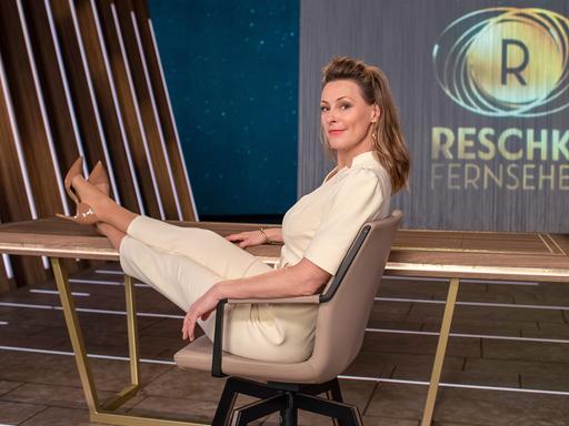 Anja Reschke sitzt in einem Bürostuhl mit den Füßen auf dem Tisch. Im Hintergrund zeigt ein Screen das Sendelogo für "Reschke Fernsehen".