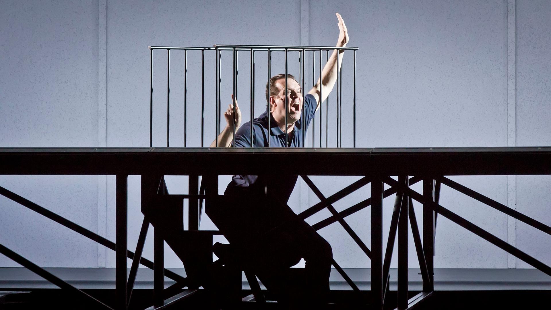 In der Inszenierung von "M - Eine Stadt sucht einen Mörder" steht der Protagonist in einer Bühnenvorrichtung, die aussieht, als befände er sich in einer Zelle. Er winkt aufgeregt.