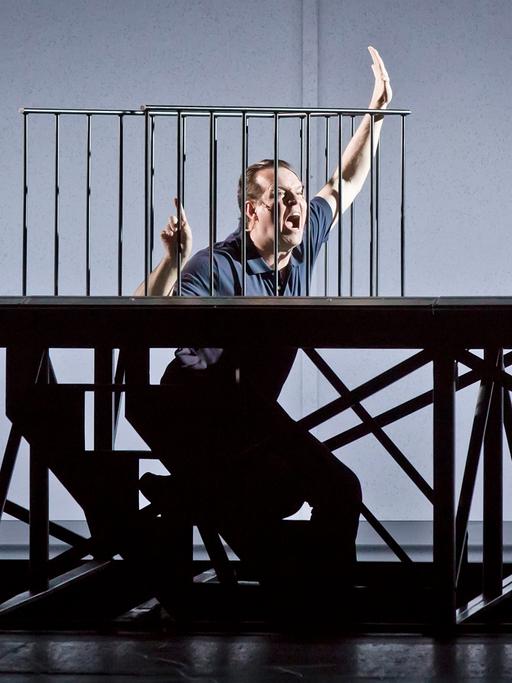 In der Inszenierung von "M - Eine Stadt sucht einen Mörder" steht der Protagonist in einer Bühnenvorrichtung, die aussieht, als befände er sich in einer Zelle. Er winkt aufgeregt.