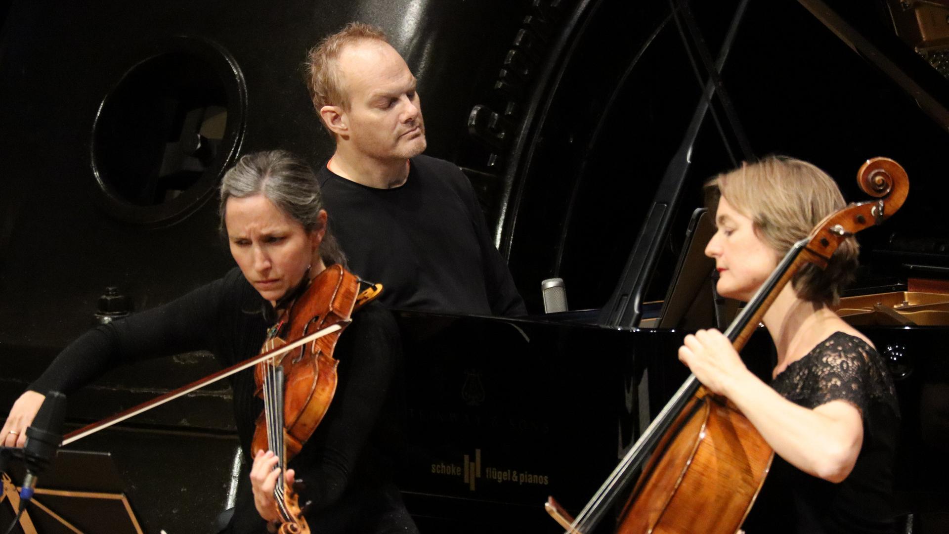 Eine Bratschistin, ein Pianist und eine Cellistin musizieren auf einer Bühne. Im Hintergrund sind Maschinen des Jugendstil-Kraftwerks Heimbach (Eifel) zu sehen. Eine Bratschistin, ein Pianist und eine Cellistin musizieren auf einer Bühne. Im Hintergrund sind Maschinen des Jugendstil-Kraftwerks Heimbach (Eifel) zu sehen.