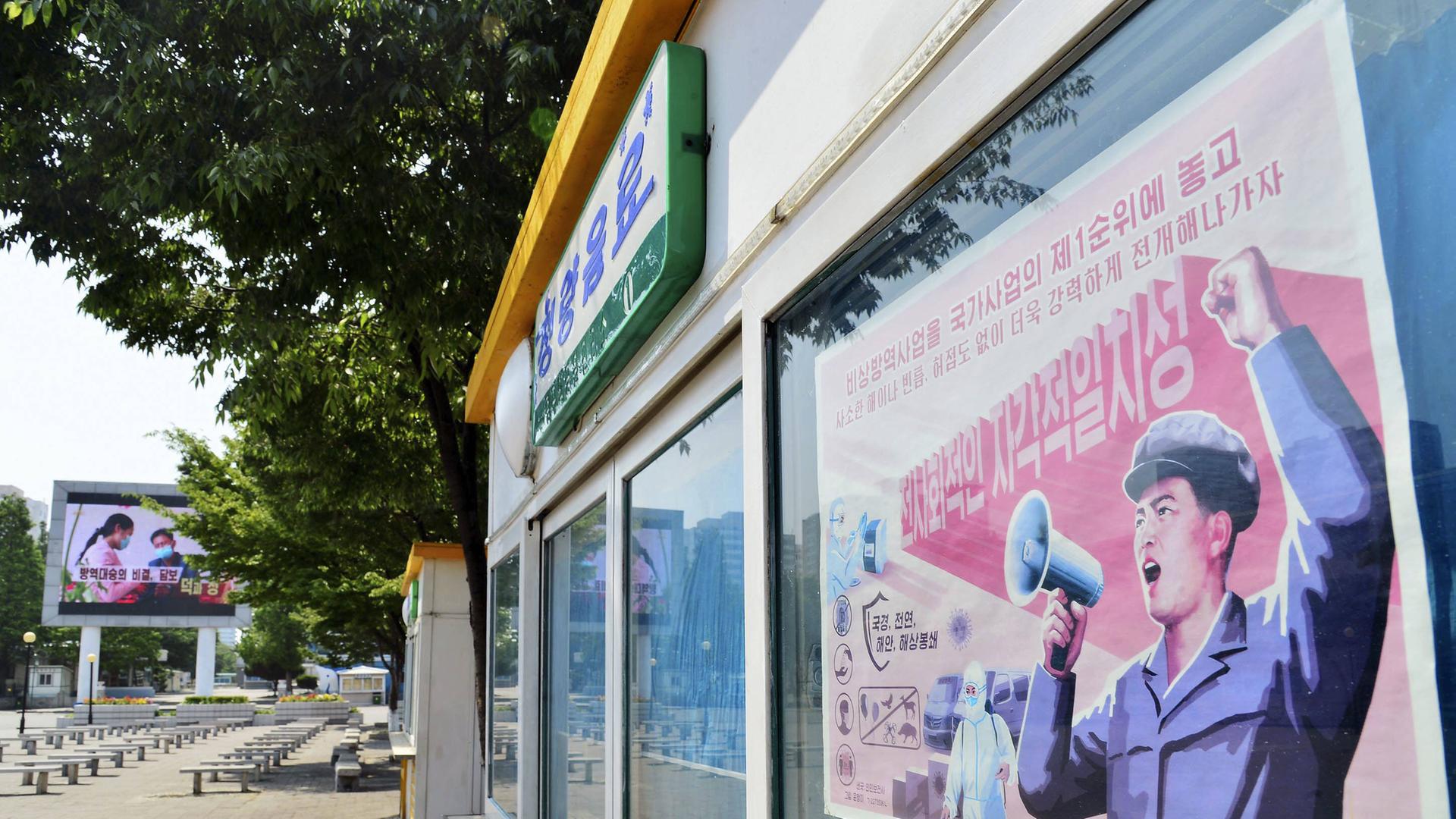 Eine leere Straße in Pjöngjang, am rechten Bildrand ein Plakat, auf dem ein Staatsbediensteter in ein Megaphon redet, neben ihm ein Mann in Schutzanzug und Maske.