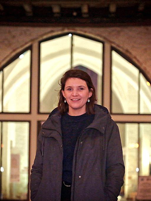 Verena Keßler steht in  dunkler Jacke vor einem großen erleuchteten Fenster.