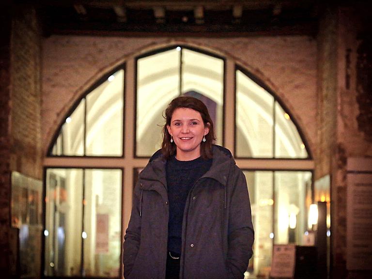 Verena Keßler steht in  dunkler Jacke vor einem großen erleuchteten Fenster.