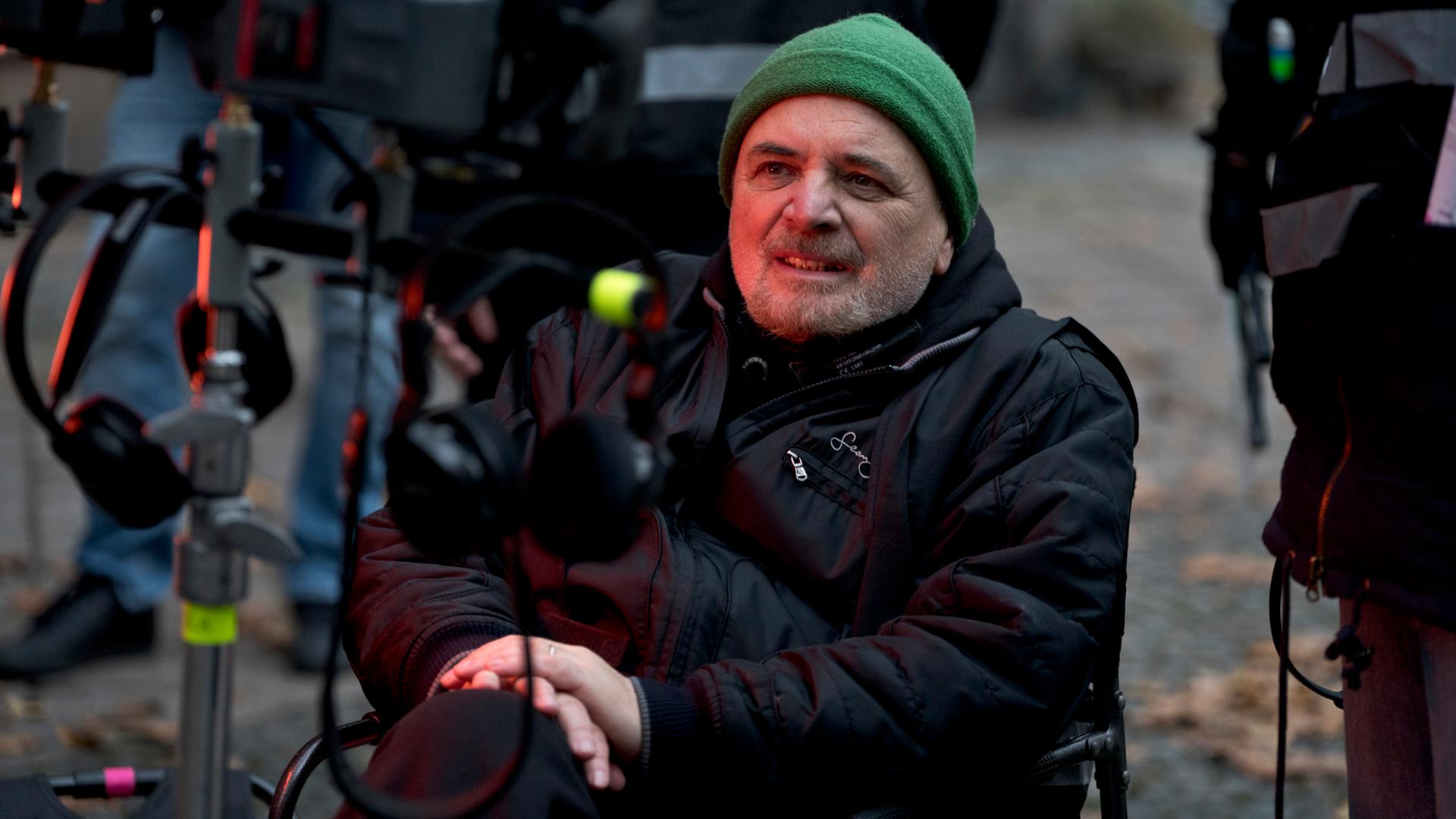 Porträt von Regisseur Uli Edel an einem Filmset. Er sitzt auf einem Stuhl und schaut auf einen Bildschirm. Er trägt eine schwarze Daunenjacke, eine grüne Mütze und einen Drei-Tage-Bart.