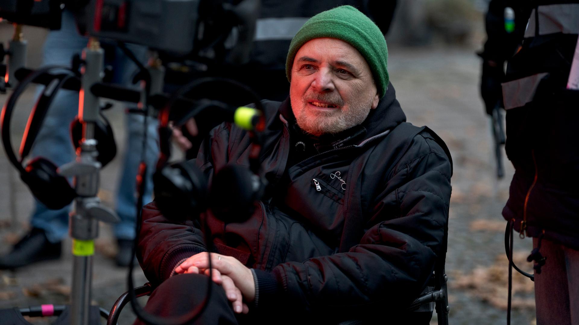 Porträt von Regisseur Uli Edel an einem Filmset. Er sitzt auf einem Stuhl und schaut auf einen Bildschirm. Er trägt eine schwarze Daunenjacke, eine grüne Mütze und einen Drei-Tage-Bart. Porträt von Regisseur Uli Edel an einem Filmset. Er sitzt auf einem Stuhl und schaut auf einen Bildschirm. Er trägt eine schwarze Daunenjacke, eine grüne Mütze und einen Drei-Tage-Bart.
