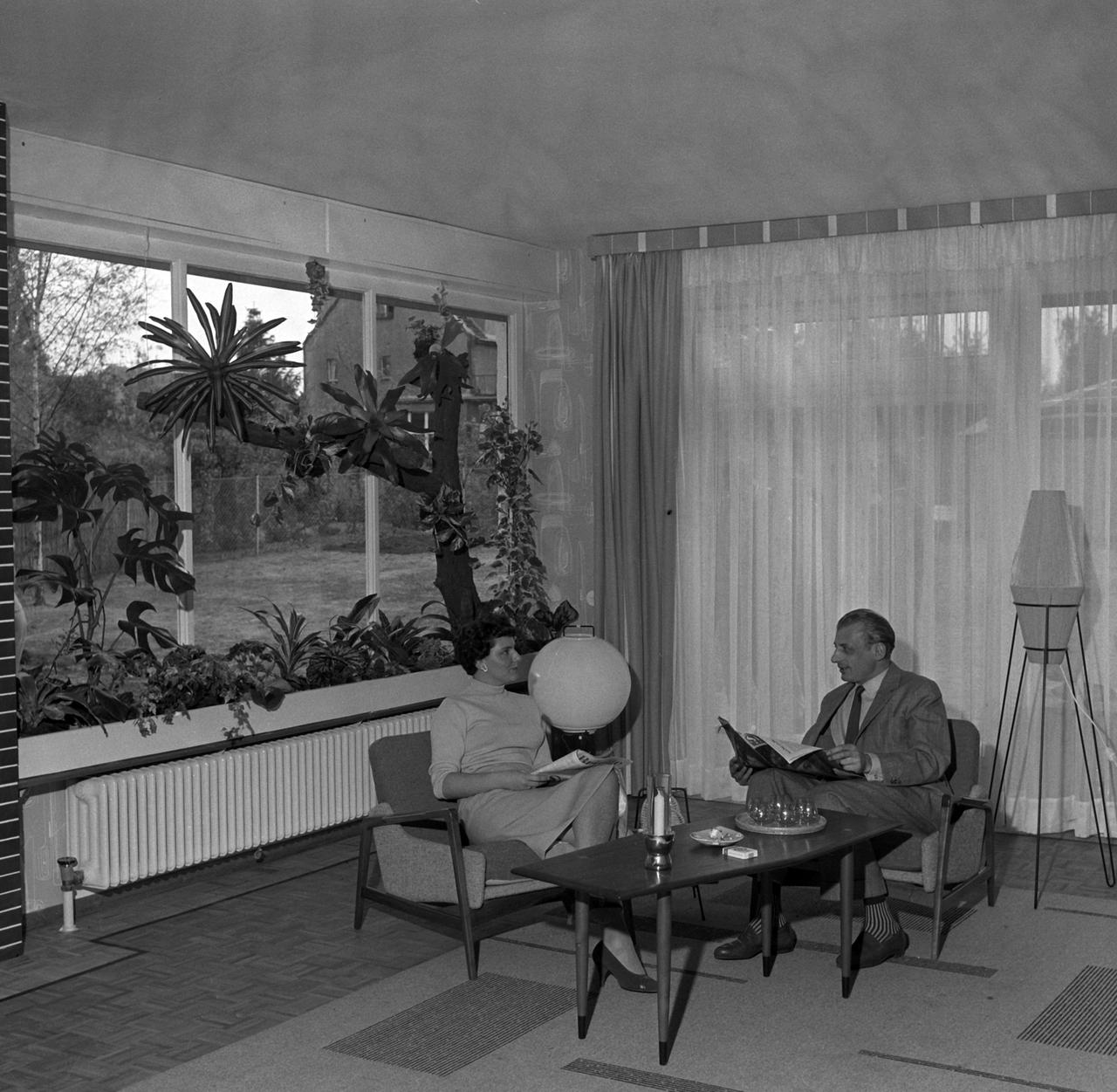 Schwarzweißfoto eines elegant gekleideten Ehepaares, das in den 1950er Jahren im Wohnzimmer eines Einfamilienhauses sitzt.