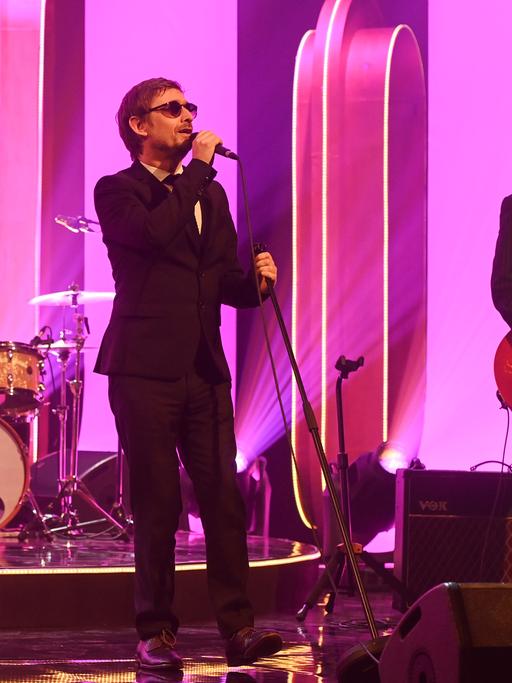 Auf einer pink-violett beleuchteten Bühne stehen vier Musikerinnen und Musiker. 