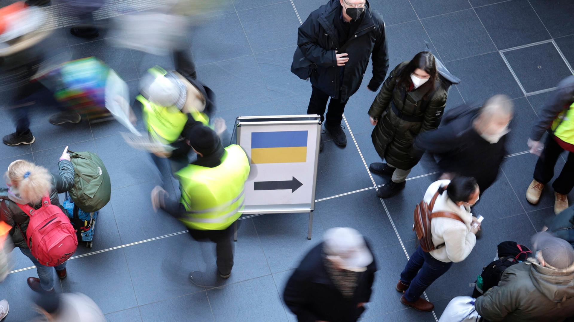 Eine Szene am Berliner Hauptbahnhof aus der Vogelperspektive: Ankunft geflüchteter Menschen aus der Ukraine am 12.3.22. Es gibt ein Hinweisschild mit der ukrainischen Flagge und einem Pfeil. Viele Menschen, manche mit Koffern, einige mit Warnwesten. Eine Szene am Berliner Hauptbahnhof aus der Vogelperspektive: Ankunft geflüchteter Menschen aus der Ukraine am 12.3.22. Es gibt ein Hinweisschild mit der ukrainischen Flagge und einem Pfeil. Viele Menschen, manche mit Koffern, einige mit Warnwesten.