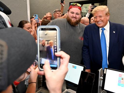 Ex-Präsident Trump und ein bärtiger Anhänger, der eine umgedrehte Baseballkappe und eine Sonnenbrille auf dem Kopf hat, lächeln in eine Smartphone Kamera.