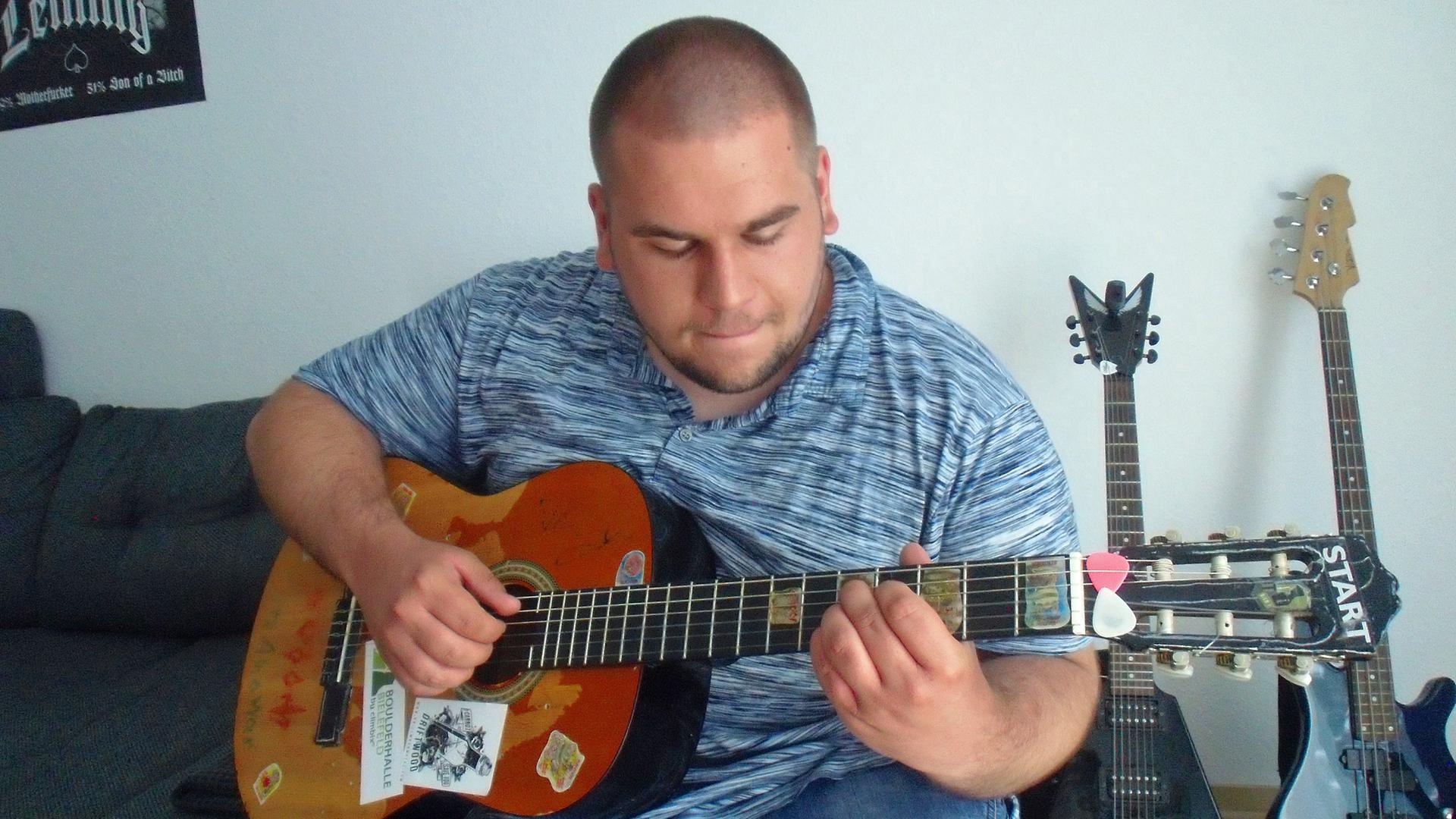 Ein junger Mann mit rasiertem Kopf spielt auf einer seiner Gitarren - weitere Instrumente sind im Hintergrund zu sehen