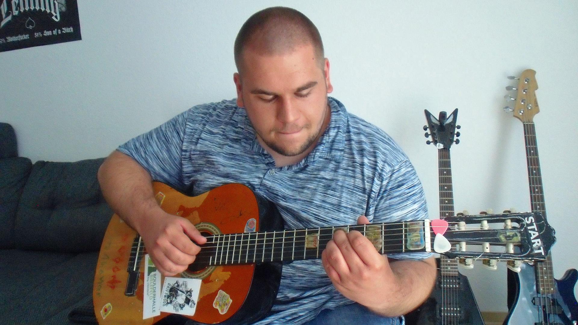 Ein junger Mann mit rasiertem Kopf spielt auf einer seiner Gitarren - weitere Instrumente sind im Hintergrund zu sehen Ein junger Mann mit rasiertem Kopf spielt auf einer seiner Gitarren - weitere Instrumente sind im Hintergrund zu sehen