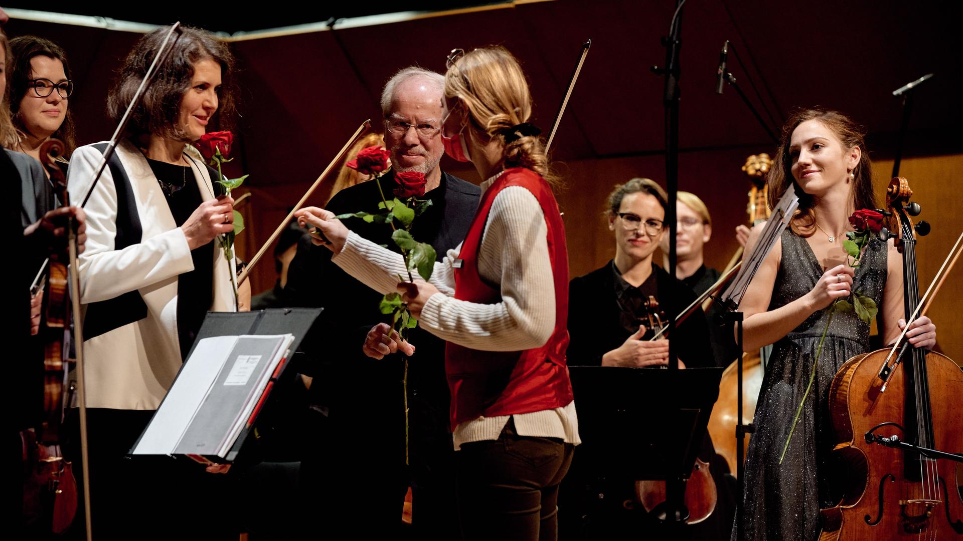 Eine rückwärtig zu sehende Frau mit Zopf verteilt Rosen an Orchester-Mitglieder, die ihre Instrumente in der Hand halten. Vor ihr stehen die Komponistin Raminta Serksnytë und der Geiger Gidon Kremer. Eine rückwärtig zu sehende Frau mit Zopf verteilt Rosen an Orchester-Mitglieder, die ihre Instrumente in der Hand halten. Vor ihr stehen die Komponistin Raminta Serksnytë und der Geiger Gidon Kremer.