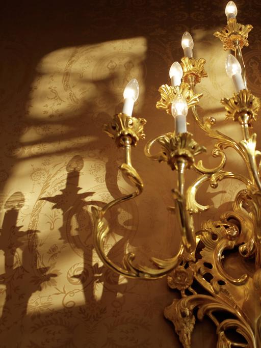 Ein goldener, barocker Wandleuchter hängt an der Wand und wirft neben ihm verschnörkelte Schatten.