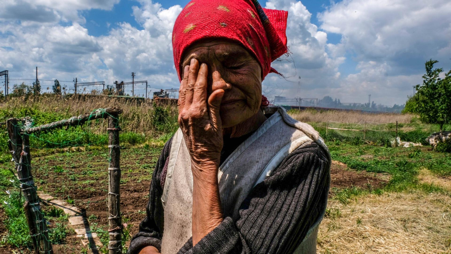 Eine alte Frau steht auf einem Feld, im Hintergrund qualmende Gebäude. Die Frau trägt ein rotes Kopftuch und wischt sich eine Träne aus dem Gesicht. 
