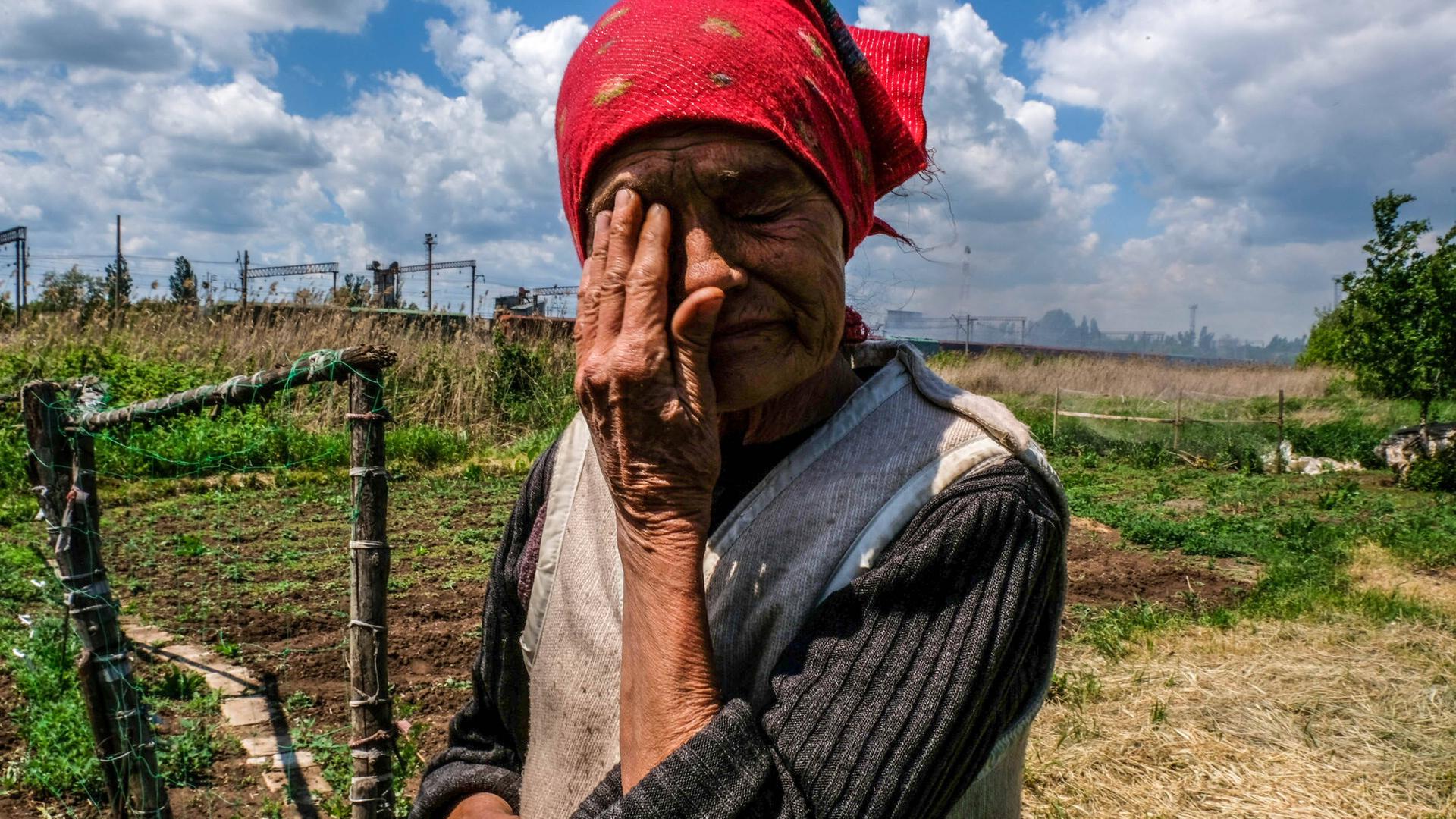 Eine alte Frau steht auf einem Feld, im Hintergrund qualmende Gebäude. Die Frau trägt ein rotes Kopftuch und wischt sich eine Träne aus dem Gesicht. Eine alte Frau steht auf einem Feld, im Hintergrund qualmende Gebäude. Die Frau trägt ein rotes Kopftuch und wischt sich eine Träne aus dem Gesicht.