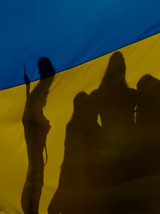 Schattenrisse von eine Gruppe von Menschen hinter einer großen blau-gelben ukrainischen Fahne. 