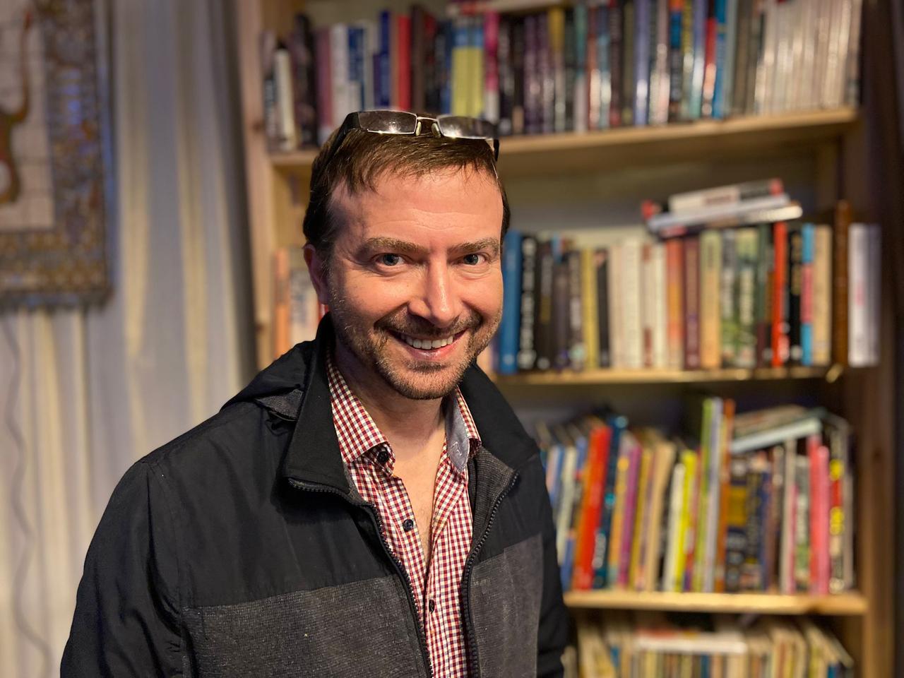 Yoni Klapholtz hat die Sonnenbrille in die Stirn geschoben und steht lächelnd vor einem Bücherregal. Yoni Klapholtz hat die Sonnenbrille in die Stirn geschoben und steht lächelnd vor einem Bücherregal.