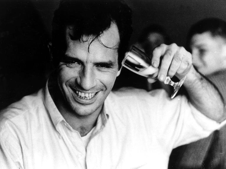 Porträt von Jack Kerouac, der sich lachend ein Sektglas ans Ohr hält.
