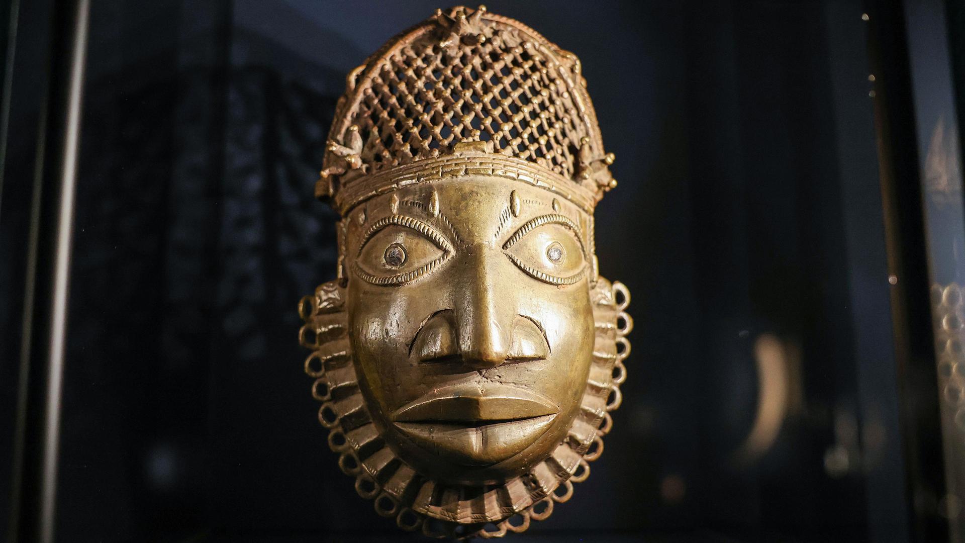 Eine golden schimmernde Maske, die das Gesicht eines Mannes darstellt, ist vor einem dunklen Hintergrund zu sehen. Eine golden schimmernde Maske, die das Gesicht eines Mannes darstellt, ist vor einem dunklen Hintergrund zu sehen.