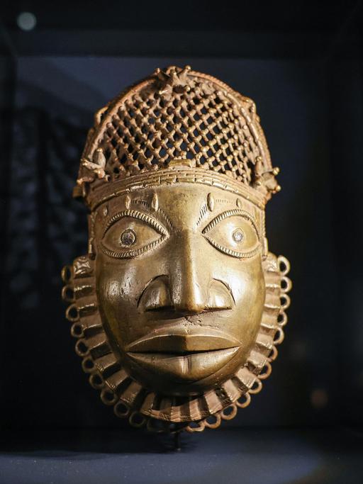 Eine golden schimmernde Maske, die das Gesicht eines Mannes darstellt, ist vor einem dunklen Hintergrund zu sehen.