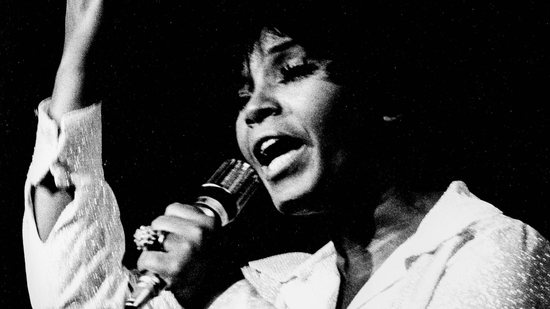 Eine schwarze Frau mit hellem Oberteil hält in der linken Hand ein Mikrofon und hebt den rechten angewinkelt an, so dass die Hand nach oben zeigt. 