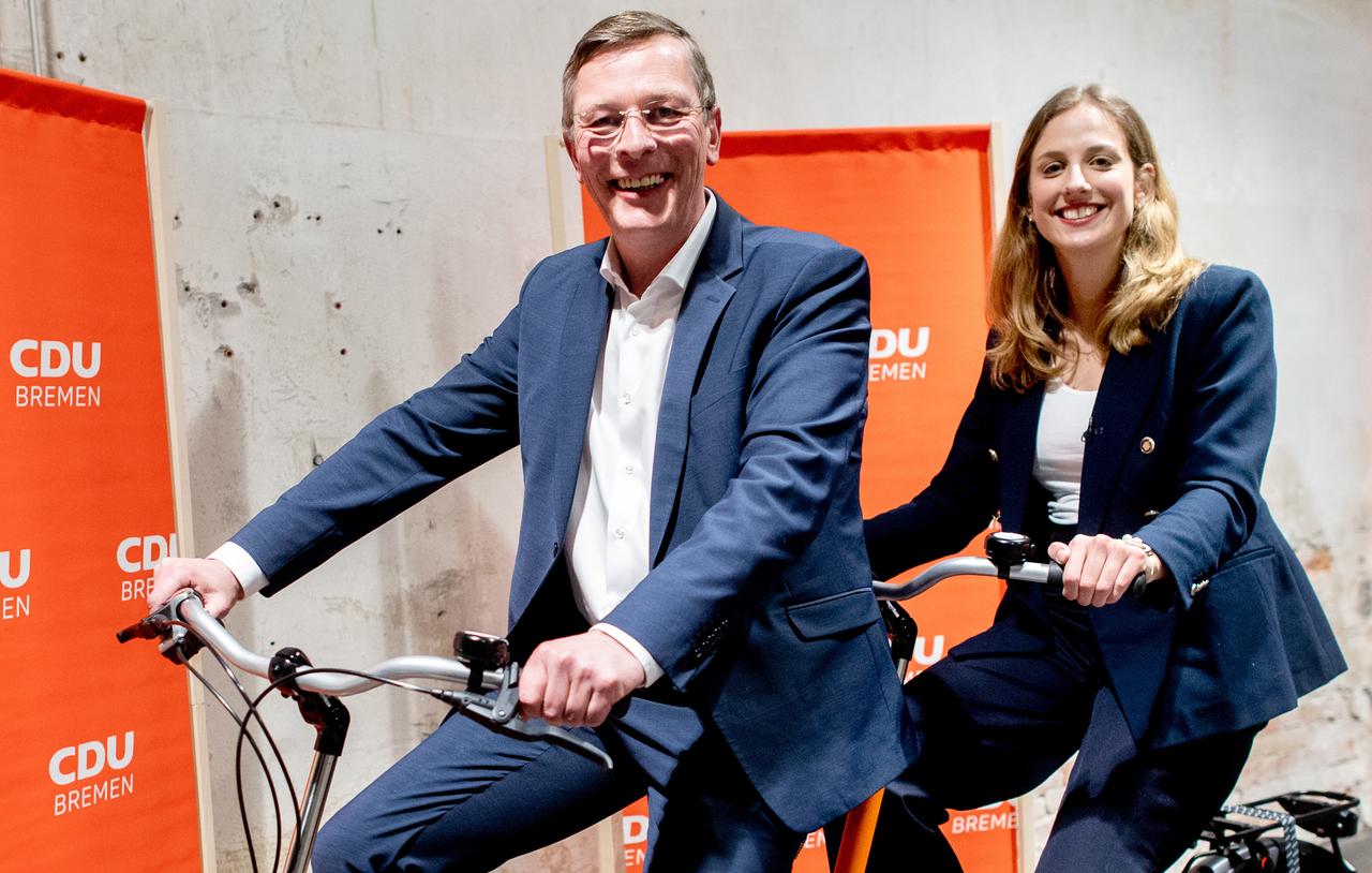 Ein älterer Mann und eine junge Frau sitzen auf einem Tandem. Hinter ihnen Wahlplakat.