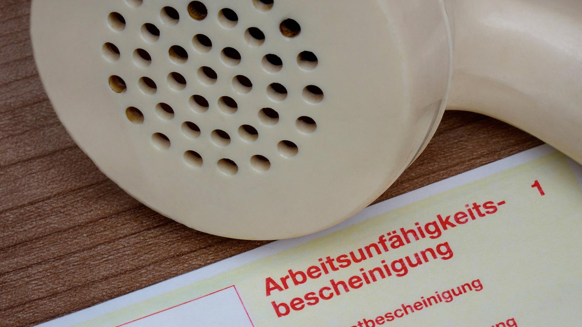 Auf einer Arbeitsunfähigkeitsbescheinigung liegt ein Telefonhörer. Auf einer Arbeitsunfähigkeitsbescheinigung liegt ein Telefonhörer.