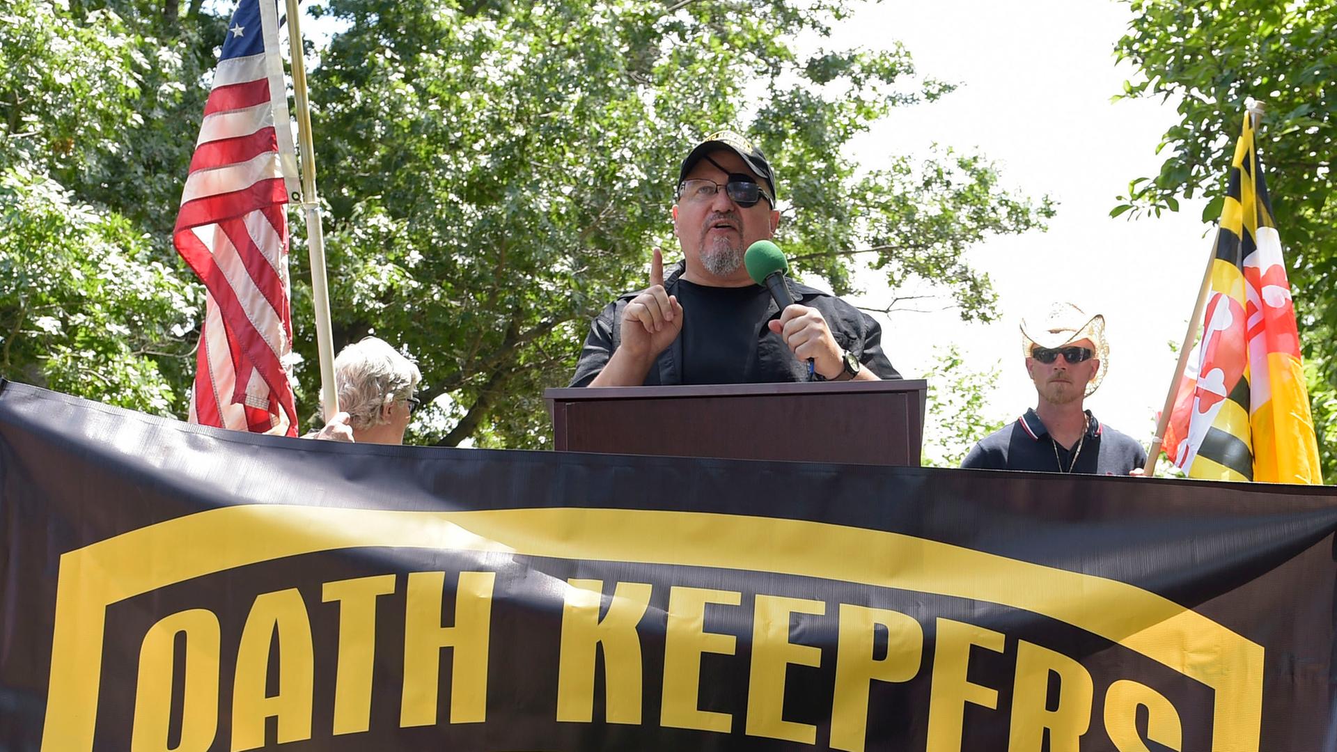 Rhodes steht an einem Redepult und spricht in ein Mikrofon. Er trägt eine Baseball-Kappe, vor ihm ist ein Banner mit der Aufschrift "Oath Keepers" aufgespannt. Im Hintergrund sieht man die US-amerikanische Flagge.