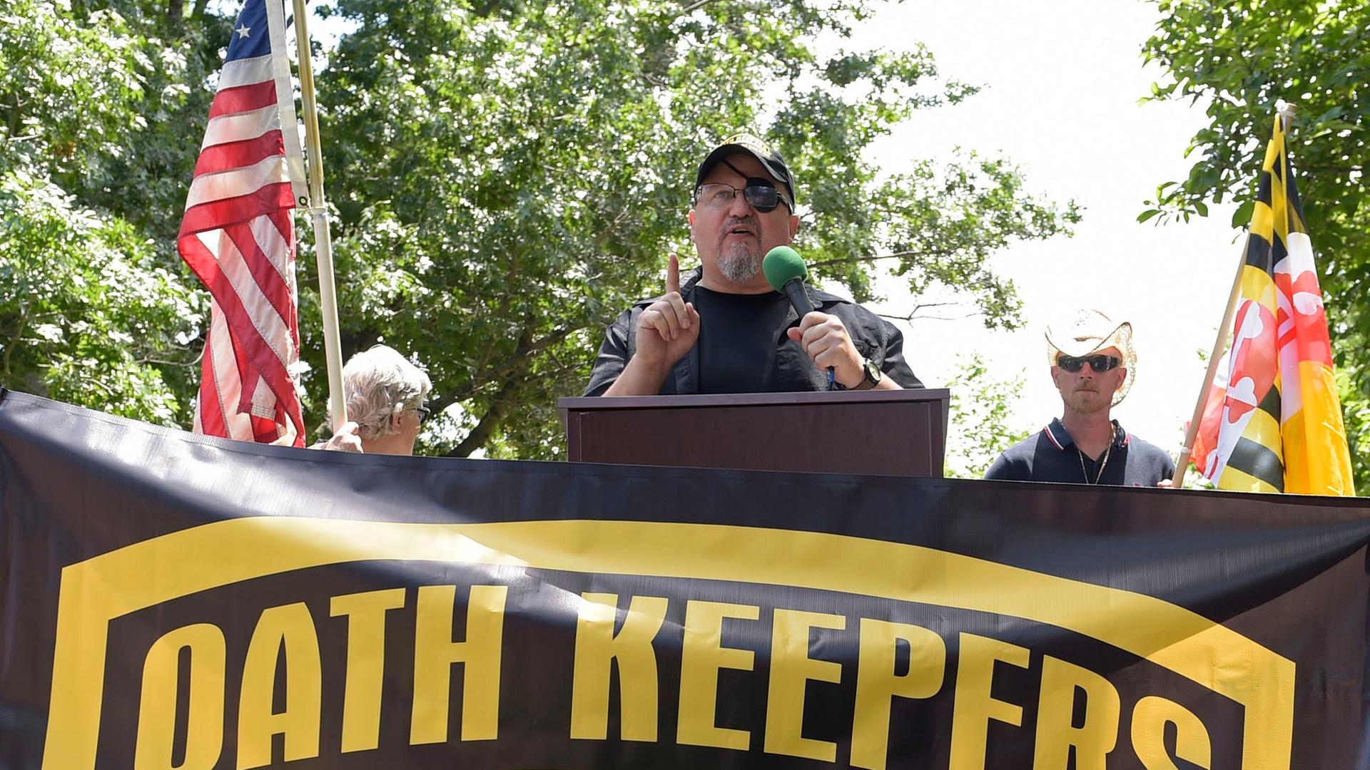 Rhodes steht an einem Redepult und spricht in ein Mikrofon. Er trägt eine Baseball-Kappe, vor ihm ist ein Banner mit der Aufschrift "Oath Keepers" aufgespannt. Im Hintergrund sieht man die US-amerikanische Flagge. Rhodes steht an einem Redepult und spricht in ein Mikrofon. Er trägt eine Baseball-Kappe, vor ihm ist ein Banner mit der Aufschrift "Oath Keepers" aufgespannt. Im Hintergrund sieht man die US-amerikanische Flagge.