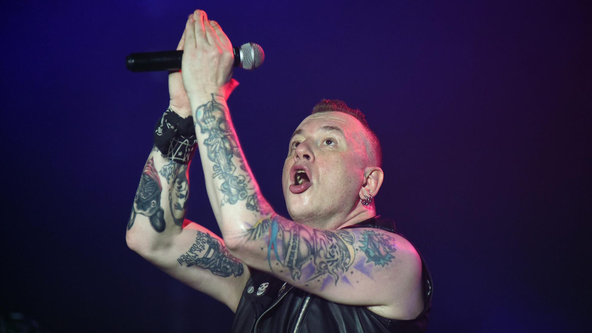 Der Sänger der Punkrockband Tarakany! singt auf einer Bühne. Er hebt seine tätowierten Arme, die das Mikrofon halten. Der Sänger der Punkrockband Tarakany! singt auf einer Bühne. Er hebt seine tätowierten Arme, die das Mikrofon halten.
