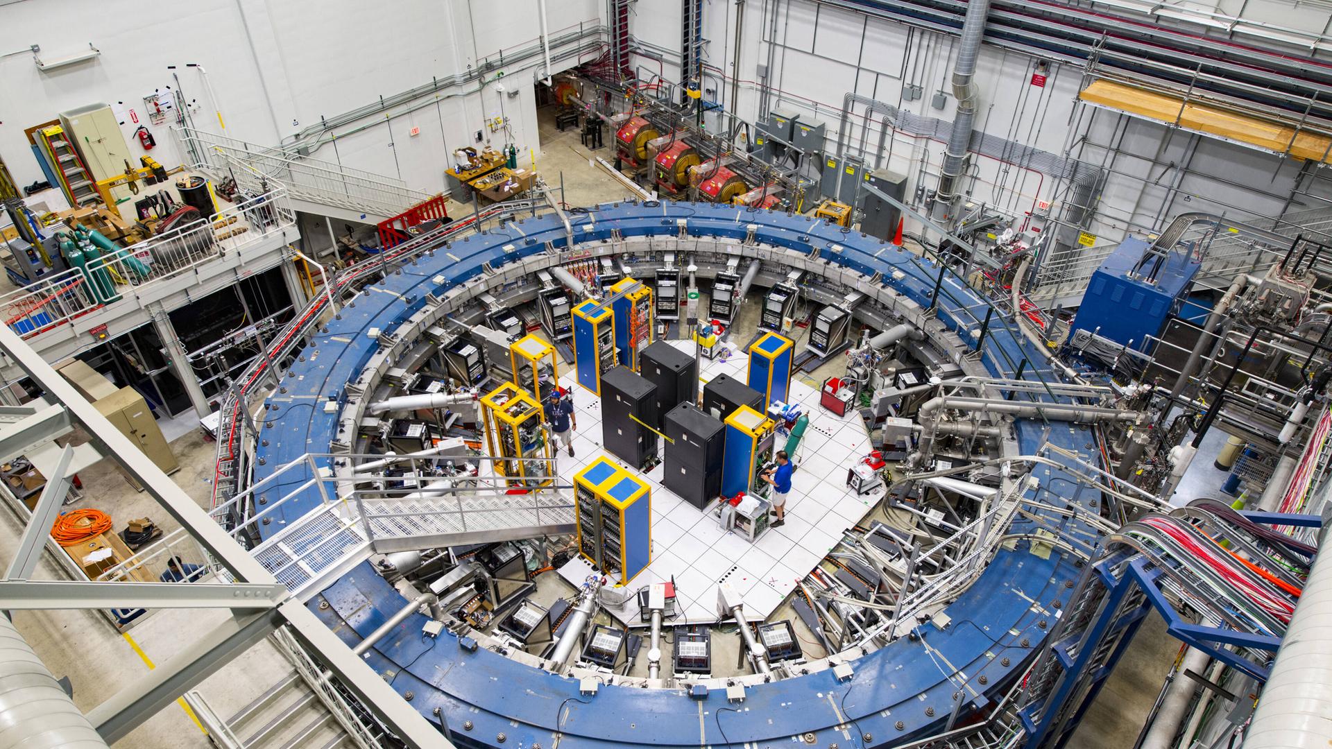 Der Teilchenbeschleuniger Myon-g-2-Ring inmitten anderer technischer Geräte in einer Halle.
Der Teilchenbeschleuniger Myon-g-2-Ring inmitten anderer technischer Geräte in einer Halle.
