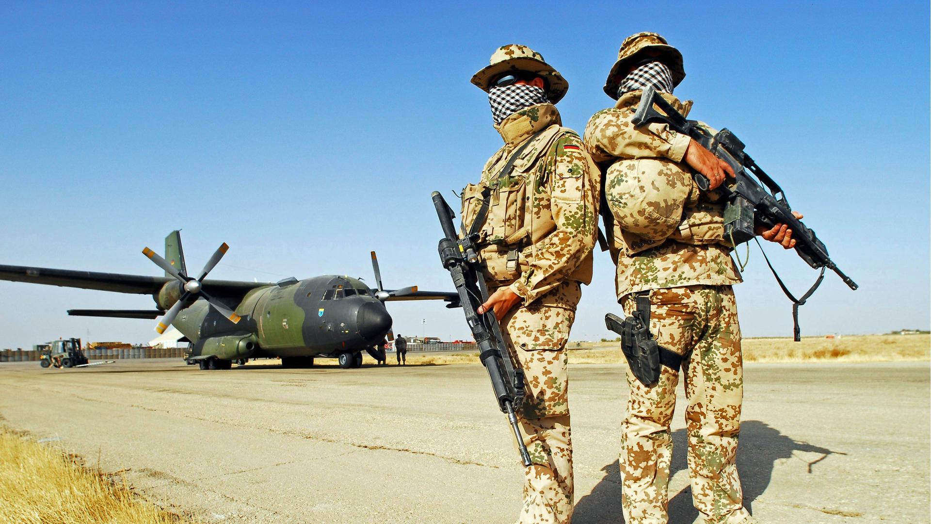 Zwei Soldaten der Bundeswehr stehen vermummt in einer Wüstenlandschaft. Im Hintergrund ein Transportflugzeug. Zwei Soldaten der Bundeswehr stehen vermummt in einer Wüstenlandschaft. Im Hintergrund ein Transportflugzeug.