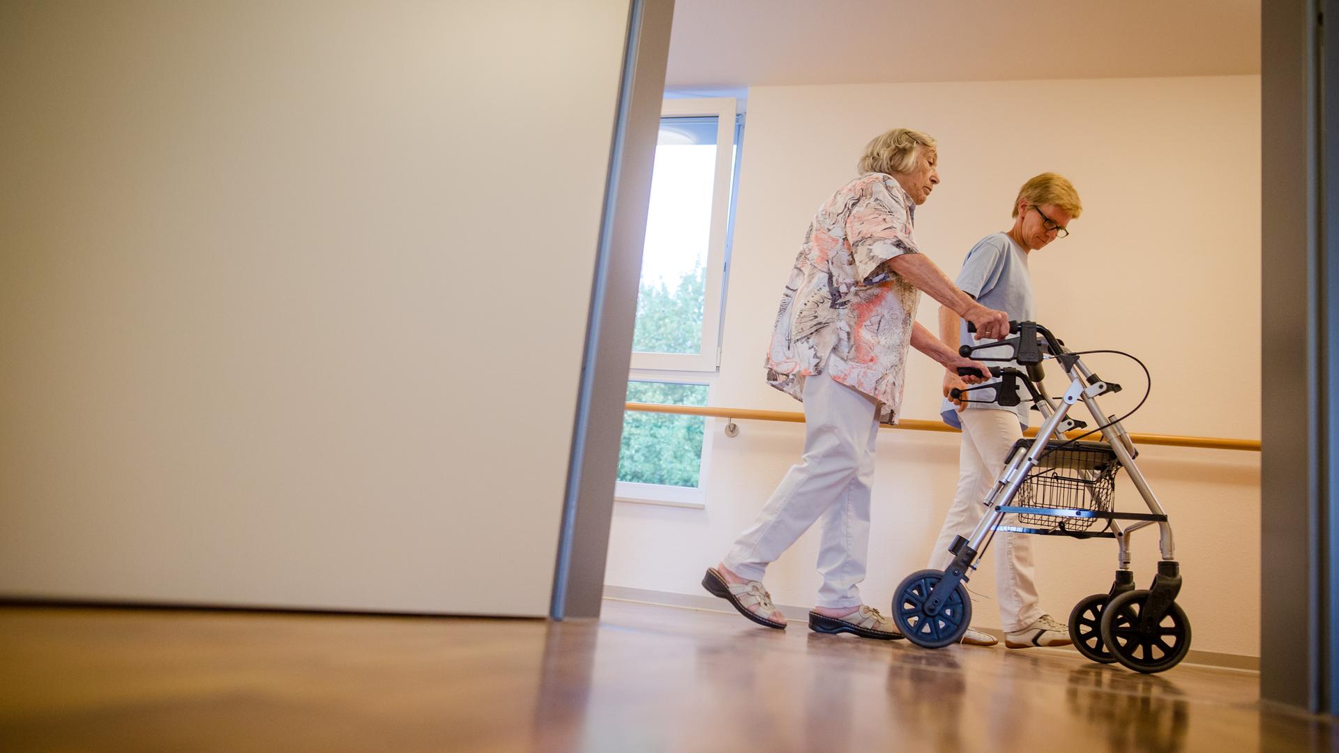 Debatte über Fachkräftemangel in der Pflege: Eine Pflegekraft geht in einem Pflegeheim mit einer älteren Dame über einen Korridor Debatte über Fachkräftemangel in der Pflege: Eine Pflegekraft geht in einem Pflegeheim mit einer älteren Dame über einen Korridor