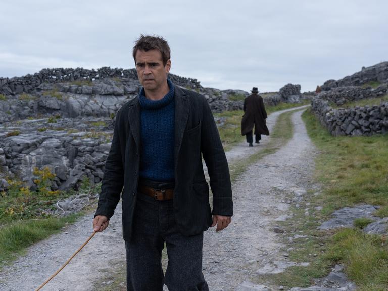 Still aus dem Film "The Banshees of Inisherin": Der Schauspieler Colin Farrell geht einen irischen Feldweg entlang. In die andere Richtung, ihm den Rücken zuwendend, geht der Schauspieler Brendan Gleeson mit Hut.