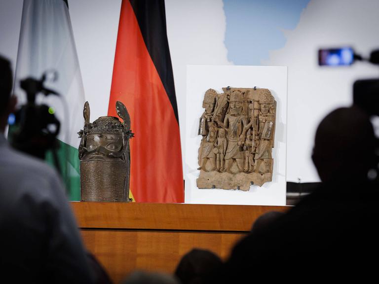 Ein verzierter Kopf aus Bronze und ein Relief mit mehrern Figuren stehen auf einem Tisch, im Hintergrund sind die Fahne Deutschlands und Nigerias. 