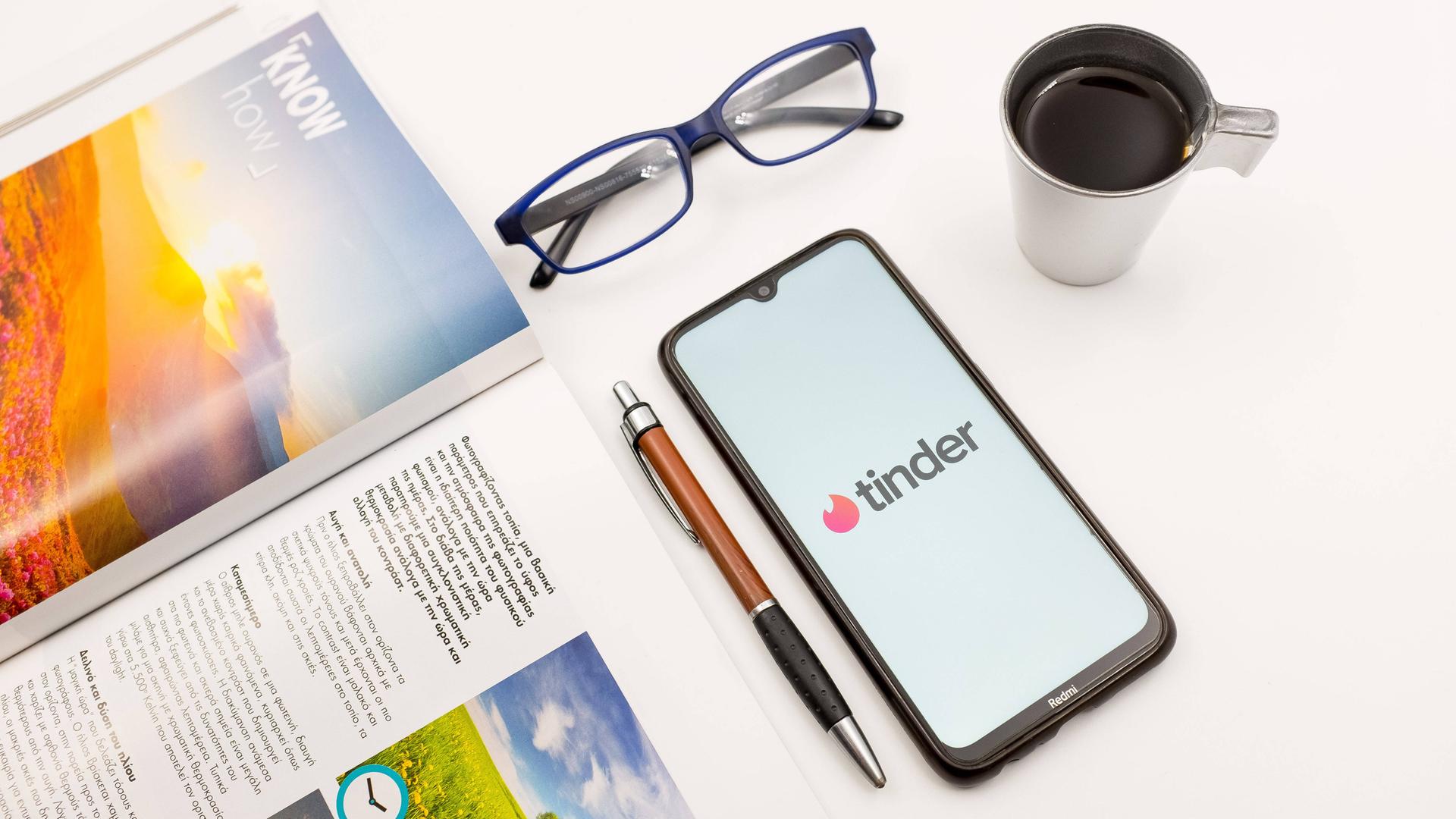 Ein Tisch mit einem Magazin, einem Stift, einer Briller und einem Smartphone auf dem Tinder zu sehen ist. Ein Tisch mit einem Magazin, einem Stift, einer Briller und einem Smartphone auf dem Tinder zu sehen ist.