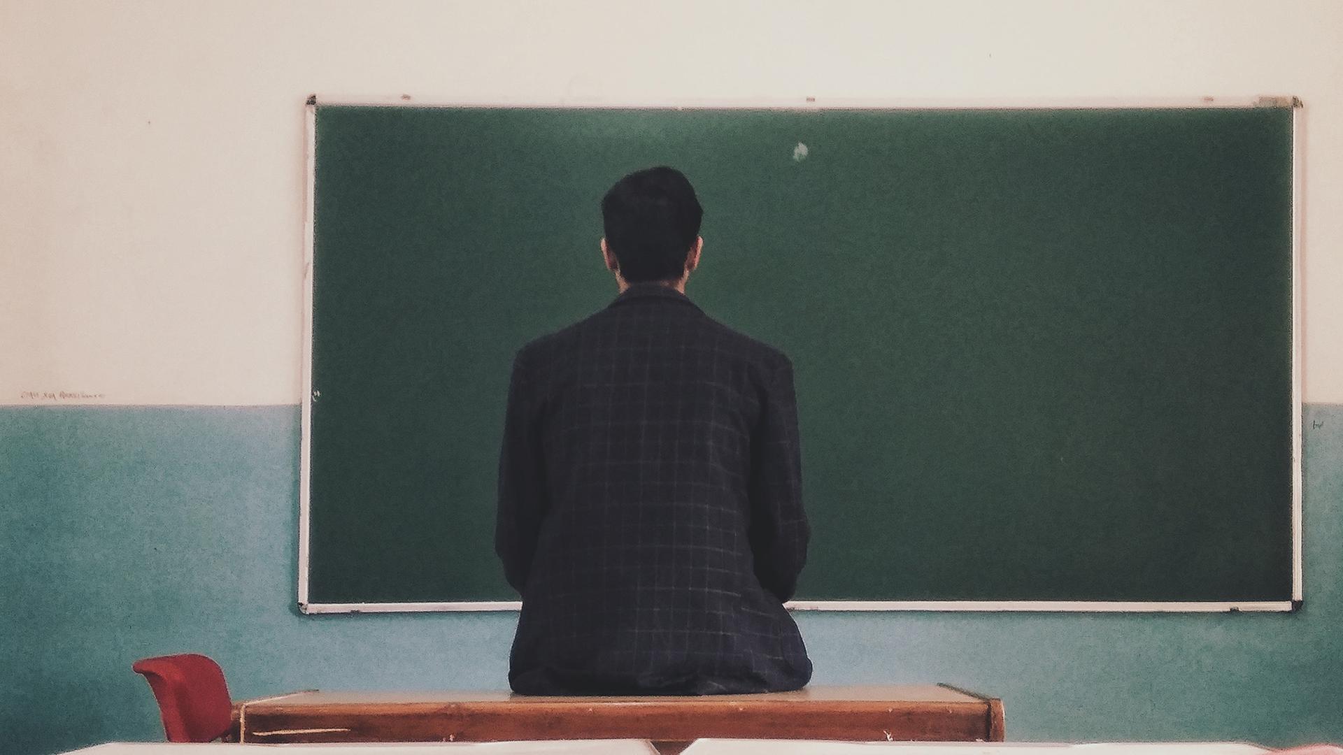 Ein Mann sitzt mit dem Rücken zur Kamera vor einer leeren Tafel in einer Schulklasse. Ein Mann sitzt mit dem Rücken zur Kamera vor einer leeren Tafel in einer Schulklasse.