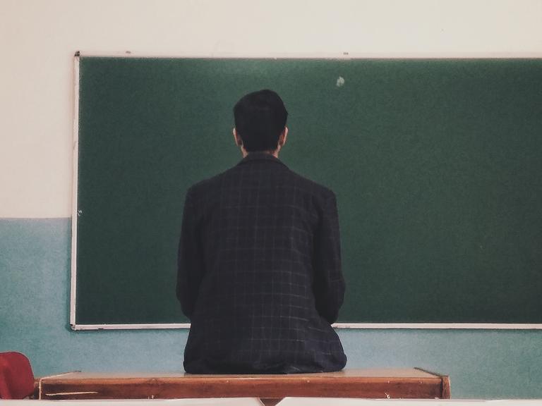 Ein Mann sitzt mit dem Rücken zur Kamera vor einer leeren Tafel in einer Schulklasse. 