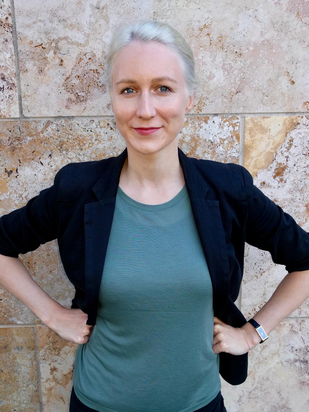 Kristina Lepold, mit zurückgekämmtem blondem Haar, einem schwarzen Sakko und grüner Bluse, hat die Hände in die Hüften gestemmt und lehnt an einer Marmorwand.