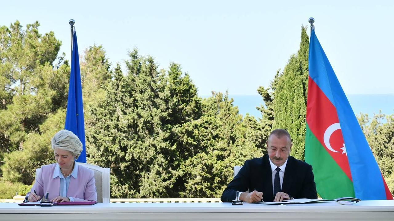 Zwei Personen, eine Frau und ein Mann, sitzen an einem langen weißen Tisch und unterzeichnen Verträge. Im Hintergrund sieht man Bäume, die Flagge der EU und von Aserbaidschan. Zwei Personen, eine Frau und ein Mann, sitzen an einem langen weißen Tisch und unterzeichnen Verträge. Im Hintergrund sieht man Bäume, die Flagge der EU und von Aserbaidschan.