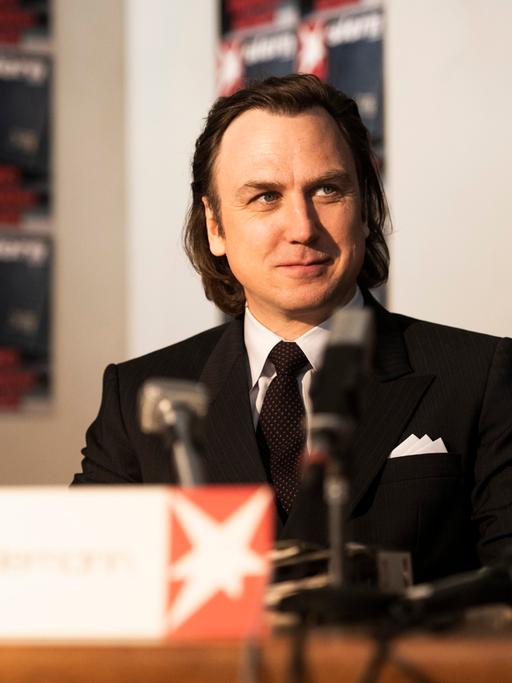 Schauspieler Lars Eidinger sitzt in seiner Figur des Reporters Gerd Heidemann in "Faking Hitler" bei einer Presskonferenz.