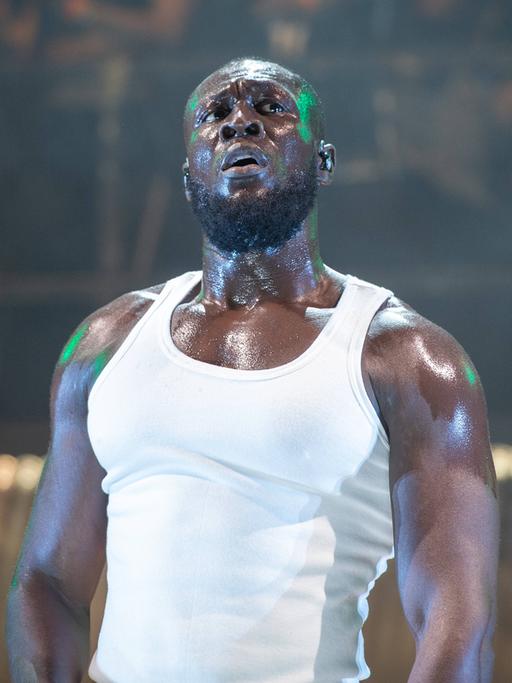 Musiker Stormzy steht im weißen Muscle-Shirt auf der Bühne. Sein Körper glänzt, Schweißperlen sind zu sehen.