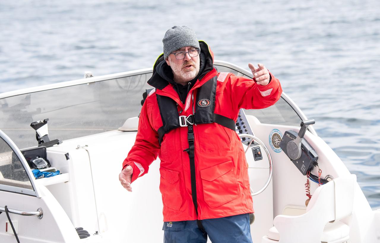 Ein Mamm in roter, wasserdichter Jacke, steht auf einem Boot und hält einen Arm auf Brusthöhe in der Luft. Er hat einen weißen Bart, trägt Brille und eine graue Wollmütze. Ein Mamm in roter, wasserdichter Jacke, steht auf einem Boot und hält einen Arm auf Brusthöhe in der Luft. Er hat einen weißen Bart, trägt Brille und eine graue Wollmütze.