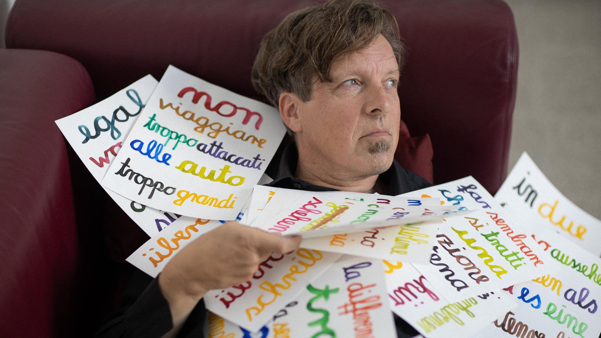 Ein Mann sitzt in einem Zugsitz und blickt zur Seite. Über seinen Oberkürper und in den Händen hält er Blätter, die mit bunter Schrift beschrieben sind. Ein Mann sitzt in einem Zugsitz und blickt zur Seite. Über seinen Oberkürper und in den Händen hält er Blätter, die mit bunter Schrift beschrieben sind.